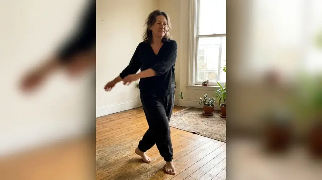 Kvinna som gungar armarna mjukt åt sidan i en vridande qigong-rörelse i ett soligt vardagsrum
